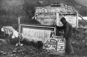 Een Tlingit-vrouw is een Chilkat-deken aan het weven, een man toont het patroonbord - Klukwan, Alaska, 1909.
