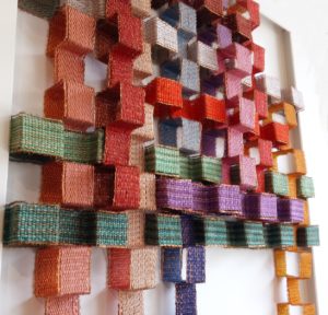 Marijke Scheepstra, Magisch vierkant. Het werkstuk is samengesteld door bandjes van 3 cm breed te weven. De weeftechniek is linnenbinding, uitgevoerd in verschillende kleuren borduurkatoen en koperdraad. Daarna heeft zij de bandjes gevouwen in blokformaat en het geheel samengevoegd tot een compositie in kleur met verschillende hoogtes.